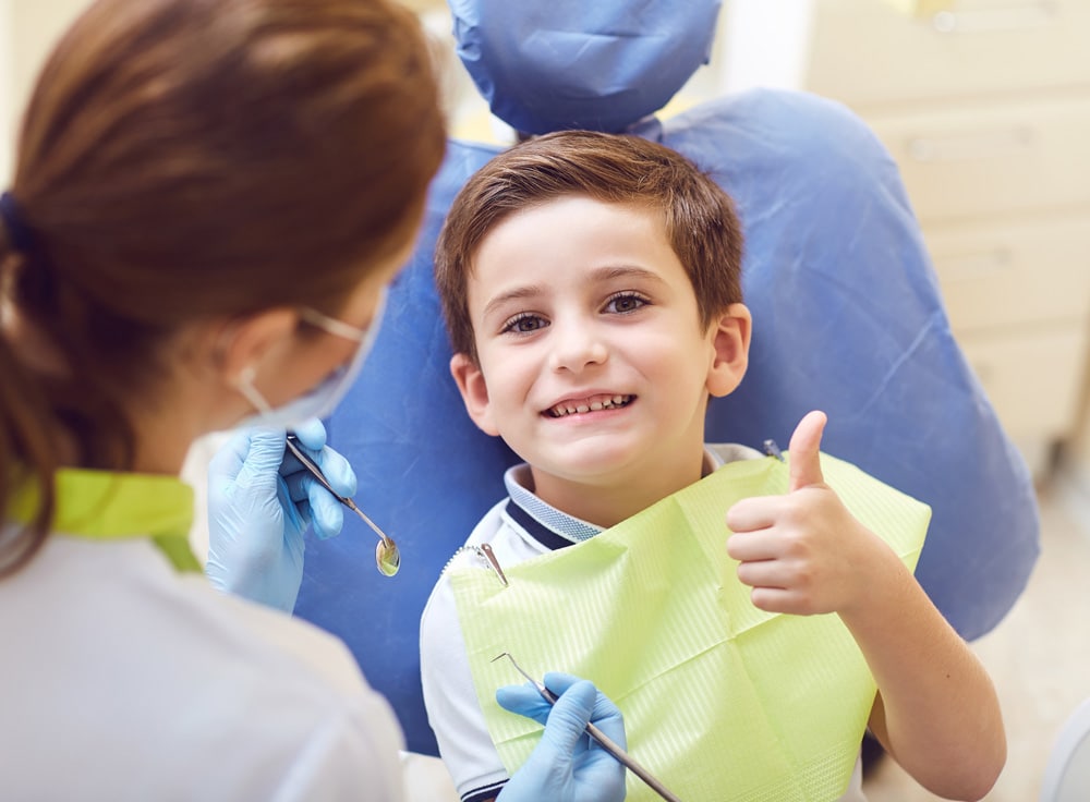 A young boy sits in a dentist chair, smiling and giving a thumbs up, showing confidence and comfort during his visit