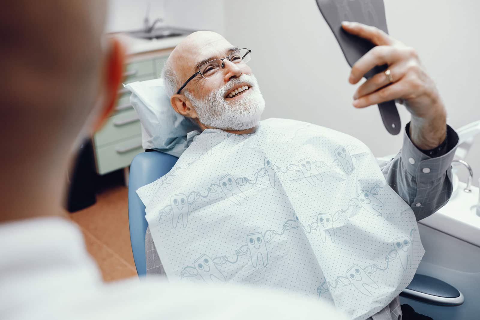 A man in a dental chair holding a mirror to examine his teeth, representing the results and care involved in reconstructive dentistry