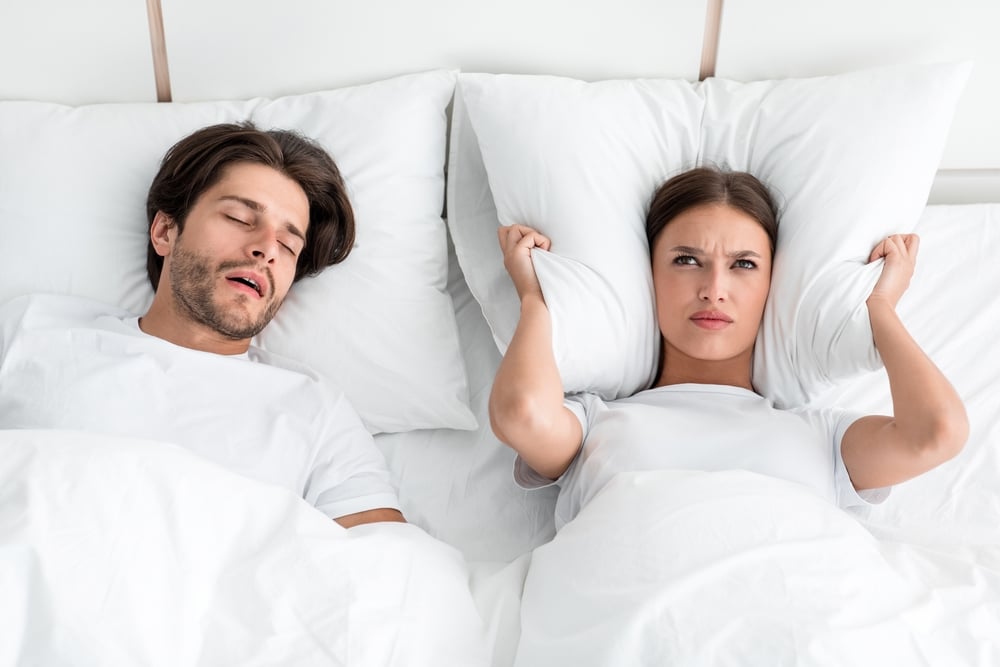 A woman covering her ears with a pillow while a man snores nearby, illustrating the disruptive effects of sleep apnea on restful sleep